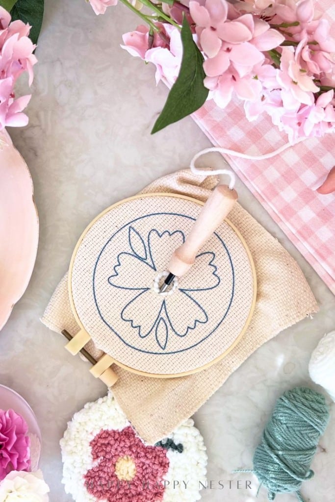 Embroidery hoop with a floral outline design on fabric, surrounded by pink flowers, green yarn for punch needle crafting DIY projects, a finished flower embroidery, and a pink gingham cloth on a light surface.