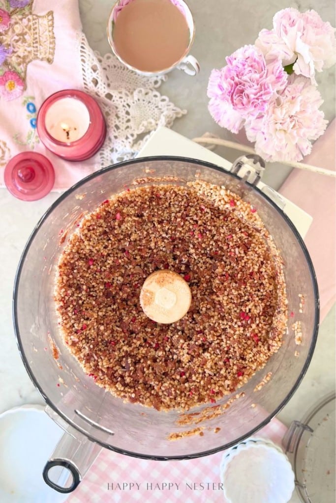 A food processor bowl filled with a crumbly nut and dried fruit mixture, inspired by our Chocolate Covered Quinoa Recipe, sits on a countertop surrounded by pink flowers, a cup of tea, a candle, and an embroidered pink napkin.