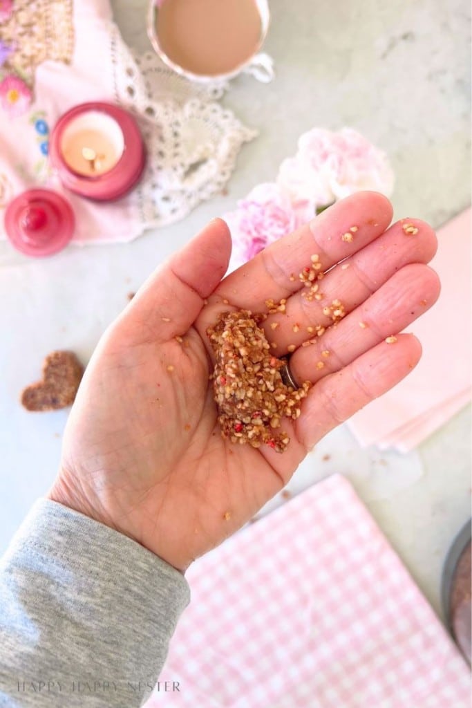 A hand holds a small piece of granola or seed snack—perhaps inspired by a Chocolate Covered Quinoa Recipe—over a light-colored table. Nearby are a pink candle, a cup of tea, a pink cloth, and a crochet doily with flowers.