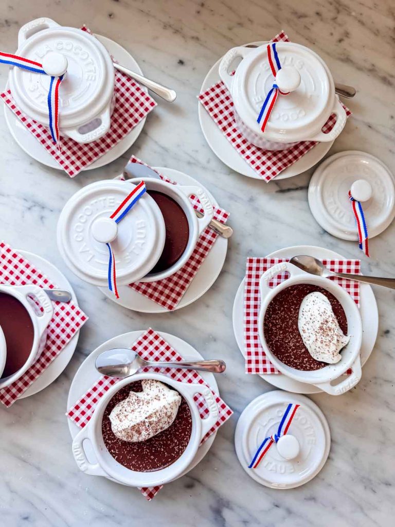 Small white ceramic pots with lids, some topped with red, white, and blue ribbons, sit on red checkered napkins. Several are open, revealing a chocolate covered quinoa recipe dessert with whipped cream on a marble surface. Spoons rest beside the pots.