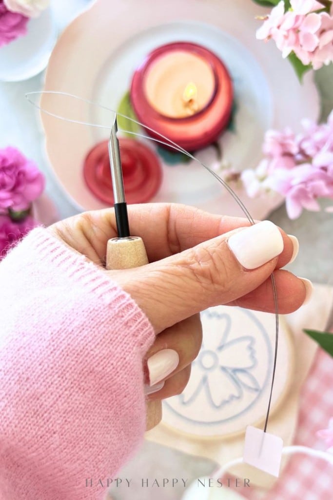 A hand with light pink nail polish holds a quilling or crafting tool with a curved wire. In the background are flowers, a red candle, and crafting materials on a pastel-colored table.