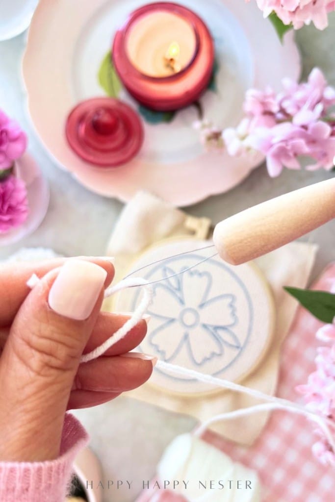 A close-up of a hand with pale pink nails holding white yarn, ready for punch needle crafting in front of a floral embroidery hoop. In the background are pink flowers, a red candle, and decorative plates—perfect for DIY inspiration.