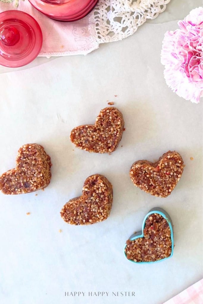 Five heart-shaped no-bake granola bars are arranged on parchment paper. One granola bar is in a blue heart-shaped cookie cutter. Pink flowers and lace are visible in the background.