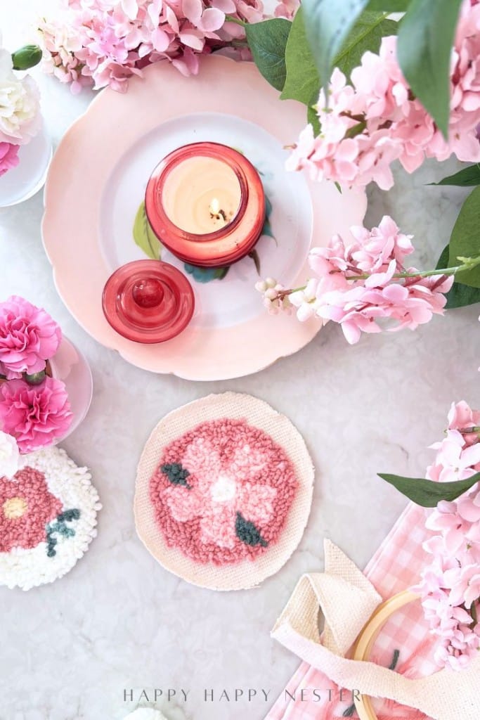 A soft pink and white table setting with pink flowers, a pink plate holding a red candle, punch needle crafting DIY coasters, and a pink gingham napkin, creating a cheerful, spring-themed arrangement.