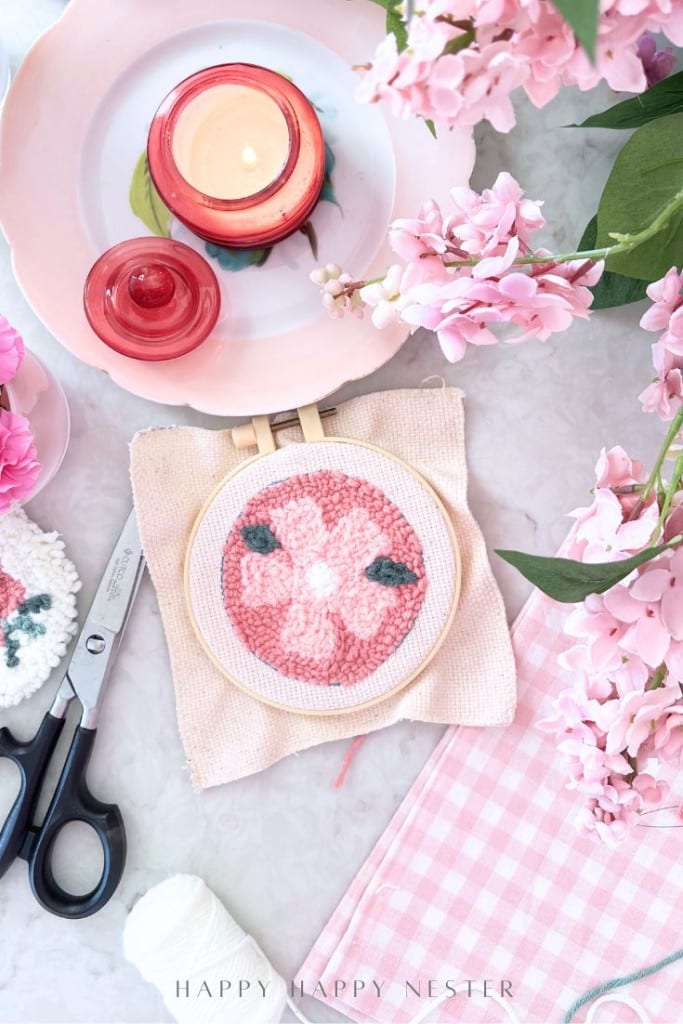 An embroidery hoop with a pink floral punch needle design sits on a table surrounded by pink flowers, a pink candle, scissors, and thread, creating a cozy, pastel punch needle crafting DIY scene.