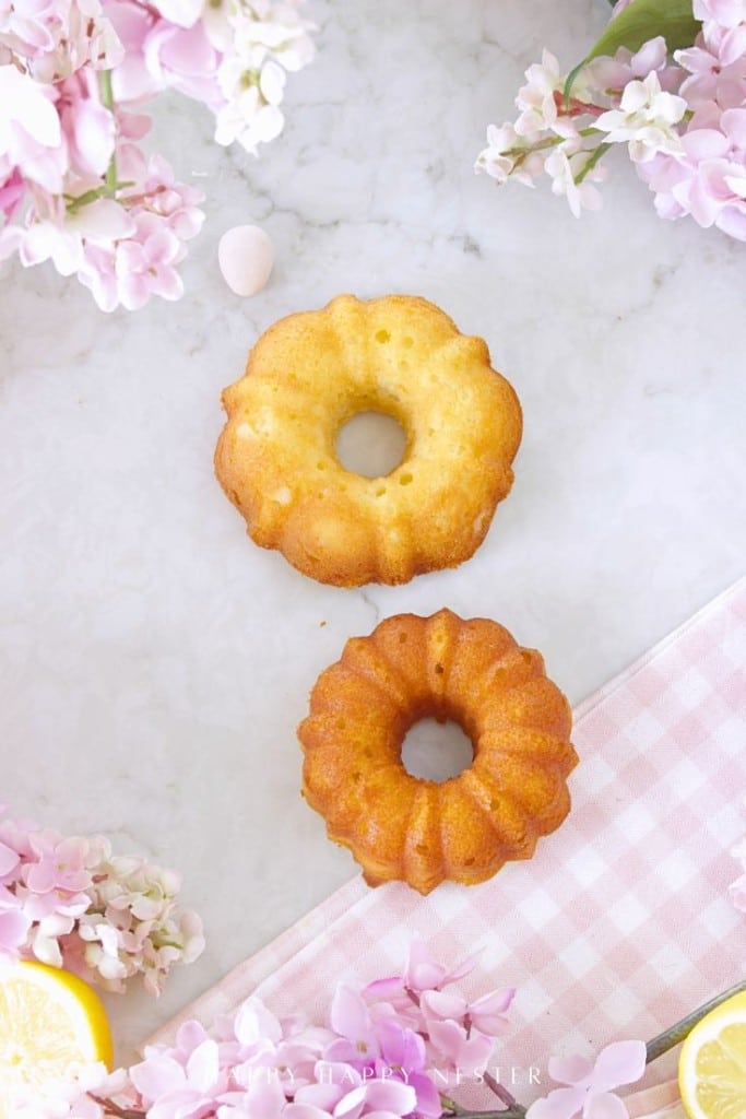 Two small bundt cakes sit on a marble surface surrounded by pink flowers, a pink gingham cloth, and lemon slices, creating a bright and spring-like setting—perfect inspiration for your Easter bundt cake recipe.