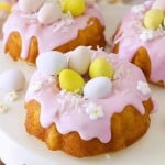 Mini bundt cakes topped with pink icing, pastel candy eggs, shredded coconut, and white flower decorations are displayed on a white cake stand—an adorable Easter bundt cake recipe perfect for your spring celebrations.