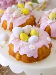 Mini bundt cakes topped with pink icing, pastel candy eggs, shredded coconut, and white flower decorations are displayed on a white cake stand—an adorable Easter bundt cake recipe perfect for your spring celebrations.