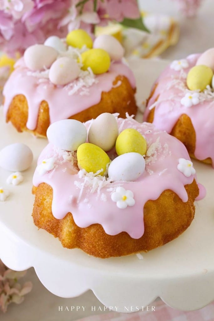 Mini bundt cakes topped with pink icing, pastel candy eggs, shredded coconut, and white flower decorations are displayed on a white cake stand—an adorable Easter bundt cake recipe perfect for your spring celebrations.