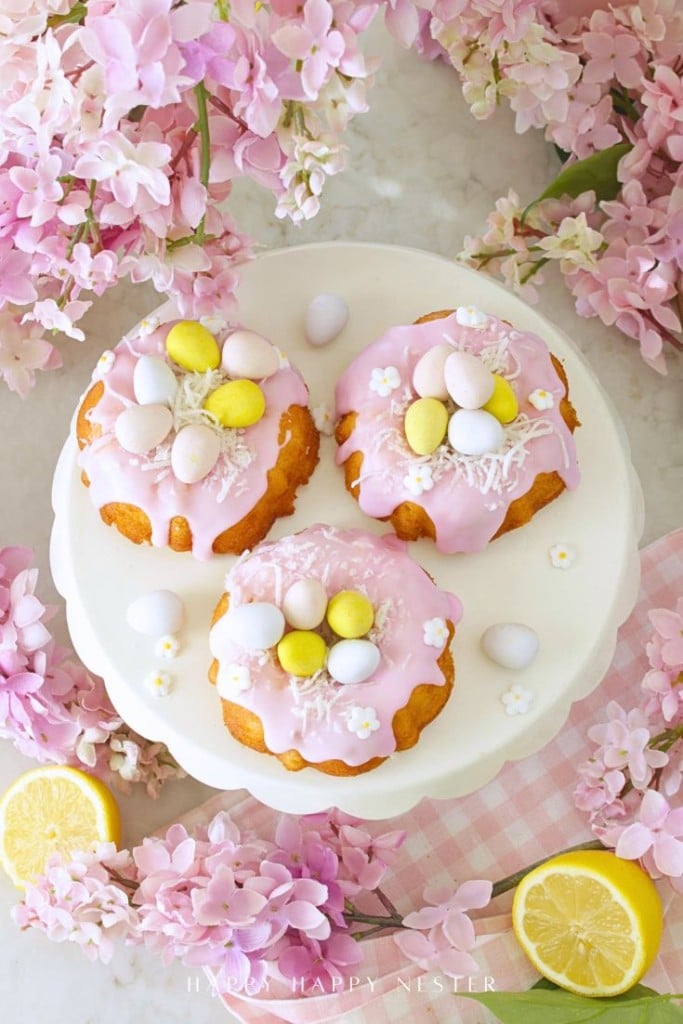 Three small bundt cakes with pink icing, decorated with pastel candy eggs and coconut flakes, sit on a white cake stand surrounded by pink flowers, lemon slices, and a pink checkered napkin.