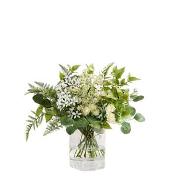 A clear glass vase filled with an arrangement of green leaves and small white and pale yellow flowers, placed against a plain white background.