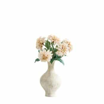 A cream-colored vase holds a bouquet of light peach dahlia flowers with green leaves, set against a plain white background.