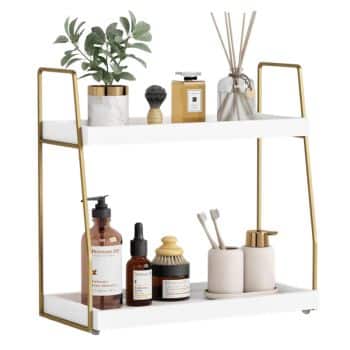 A two-tier white and gold shelf holds toiletries, including lotion, soap, toothbrushes in a cup, perfume, a plant, a candle, and reed diffusers, neatly organized for bathroom or vanity use.