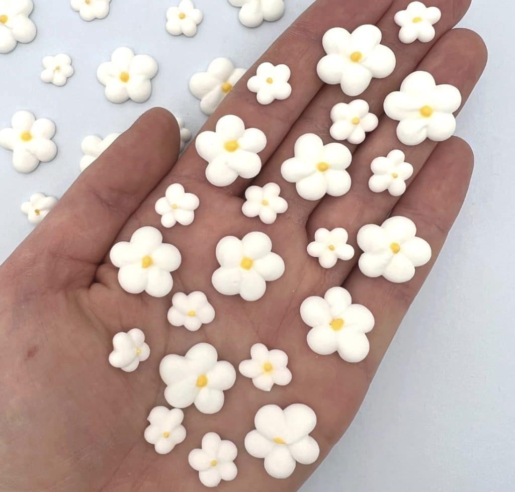 A hand displays multiple small, white, five-petaled flower-shaped decorations with yellow centers—perfect for topping an Easter bundt cake—with more scattered on a white surface.