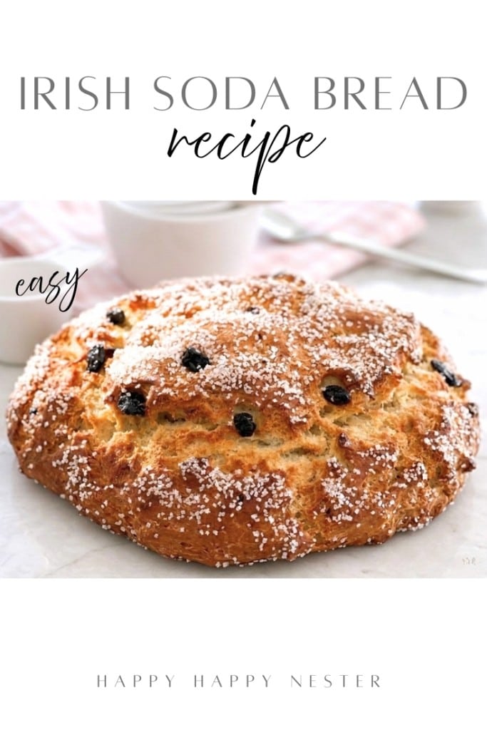 A round loaf of Irish soda bread recipe (with buttermilk) dusted with powdered sugar and dotted with raisins sits on a white surface, with cups and a pink napkin in the background. Text reads "Irish Soda Bread Recipe" and "easy.