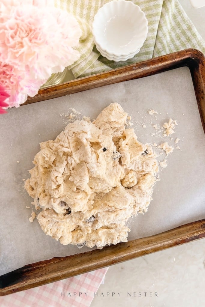 A mound of unbaked scone dough with chocolate chips sits on parchment paper on a baking sheet, surrounded by a pink flower, a green checked cloth, and small empty white bowls.