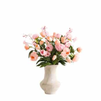 A white ceramic vase filled with an arrangement of pink and peach peonies and green leaves, set against a plain white background.