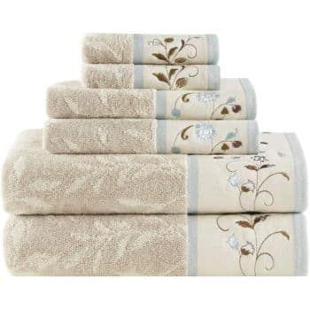 A stack of six beige towels in varying sizes, each featuring light blue trim and floral embroidery with white and brown flowers near the edge.