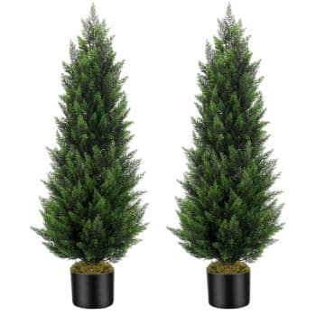 Two tall, green, artificial pine trees in black pots are placed side by side against a white background. The trees are dense with needle-like foliage and conical in shape.