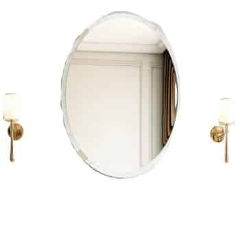 A round wall-mounted mirror with a wavy edge is flanked by two gold wall sconces with white cylindrical lampshades, reflecting part of a neutral-colored room with decorative wall panels.