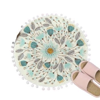 A round white rug with blue and yellow floral patterns and pom-pom edging. A pair of light pink slippers is placed on the lower right edge of the rug.