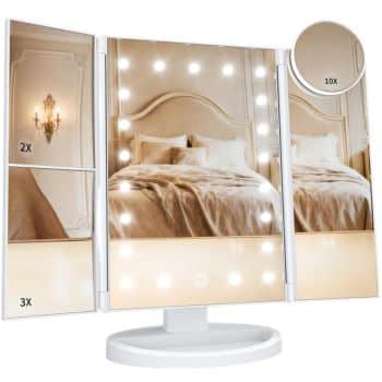 A tri-fold vanity mirror with built-in LED lights displays reflections of an elegant bedroom. The left panel has 2x and 3x magnification, and a small detachable 10x magnifying mirror is attached to the right panel.