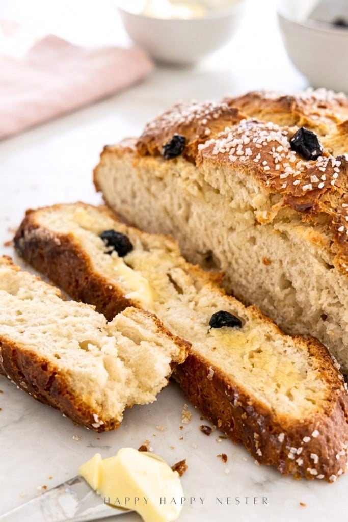 A loaf of braided bread with coarse sugar and raisins on top, partially sliced. Two slices are cut, one spread with butter. In the background, there is a butter dollop and a soft pink napkin.