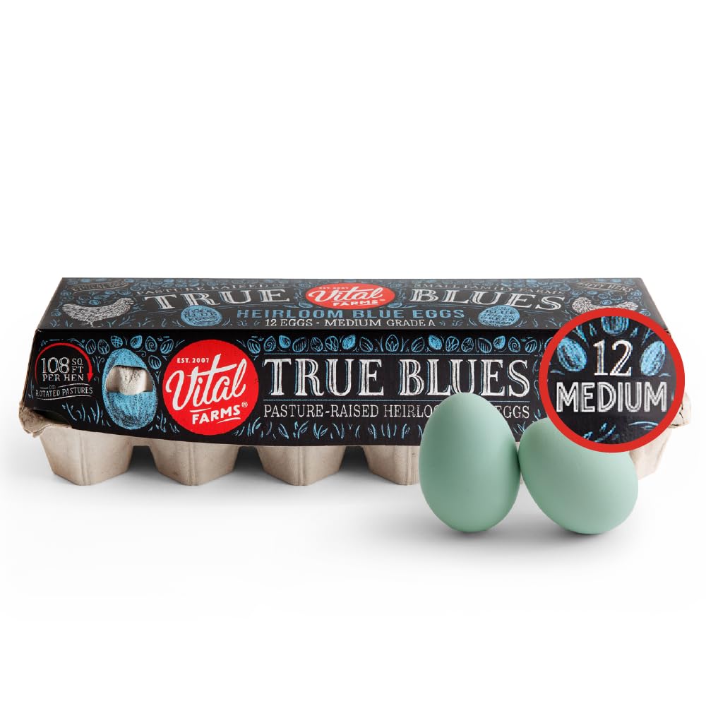 A carton of Vital Farms True Blues pasture-raised heirloom blue eggs with two light blue eggs placed in front, next to a delicate spring flower wreath. The carton shows 12 medium eggs and features black, blue, and red labeling.