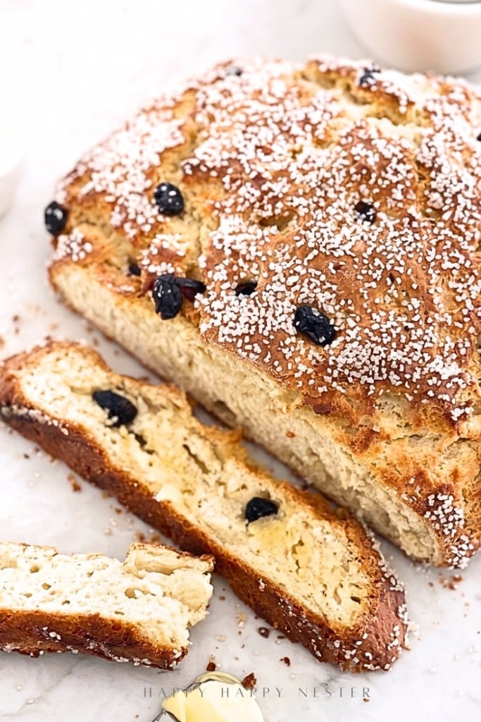 A loaf of golden-brown, sugar-topped bread with raisins is partially sliced on a white surface. Two slices are lying in front, showing the soft, airy interior and bits of raisins inside.