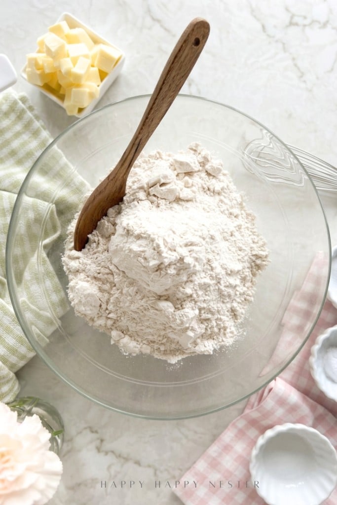 A glass bowl filled with flour and a wooden spoon sits on a marble countertop, ready for an Irish soda bread recipe (with buttermilk), surrounded by cubes of butter, a whisk, and small bowls atop pastel-colored cloths.