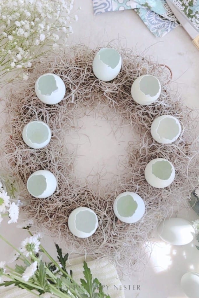 A spring flower wreath DIY features dried moss adorned with white eggshell halves, arranged in a circle. White flowers and green leaves surround the wreath, creating a soft, spring-themed display.
