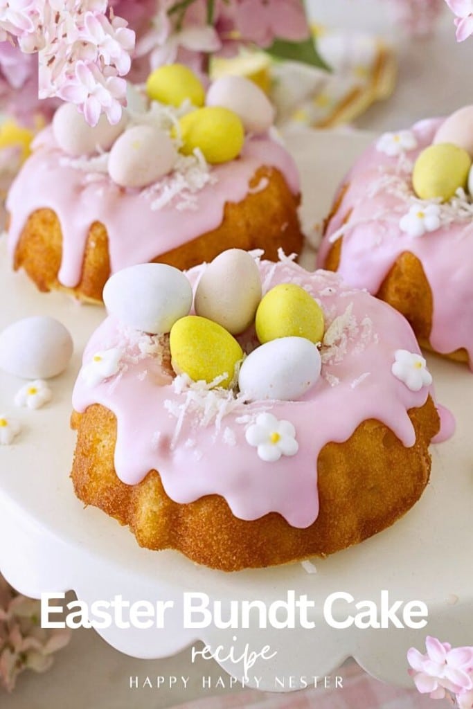 Mini bundt cakes with pink icing, decorated with pastel candy eggs, white sprinkles, and small flower shapes, displayed on a white surface with pink flowers. Text reads: “Easter bundt cake recipe.” Perfect for your festive celebrations!.