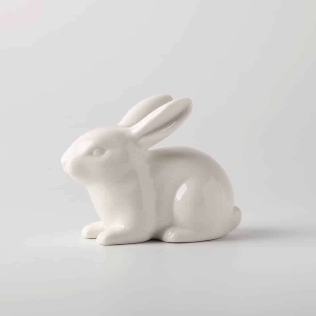 A smooth, white ceramic bunny figurine is shown against a plain light gray background. The bunny sits upright with its ears back, its glossy finish reflecting soft light—perfect for pairing with a spring flower wreath.