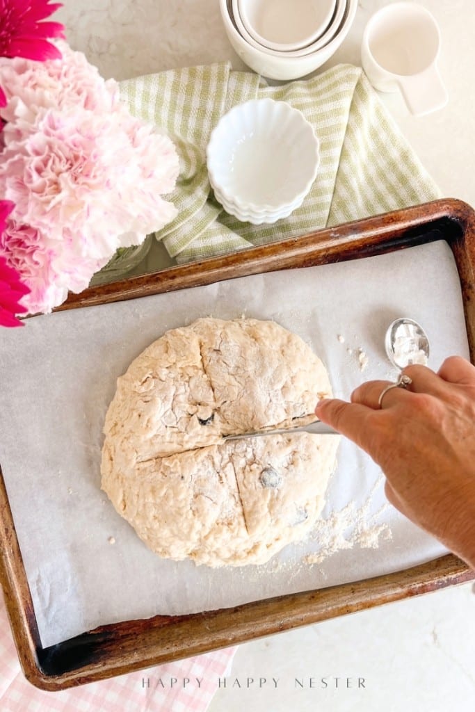 A hand uses a knife to score a round loaf of dough on a parchment-lined baking sheet. Pink flowers, stacked dishes, and a green striped cloth are nearby on a light surface.