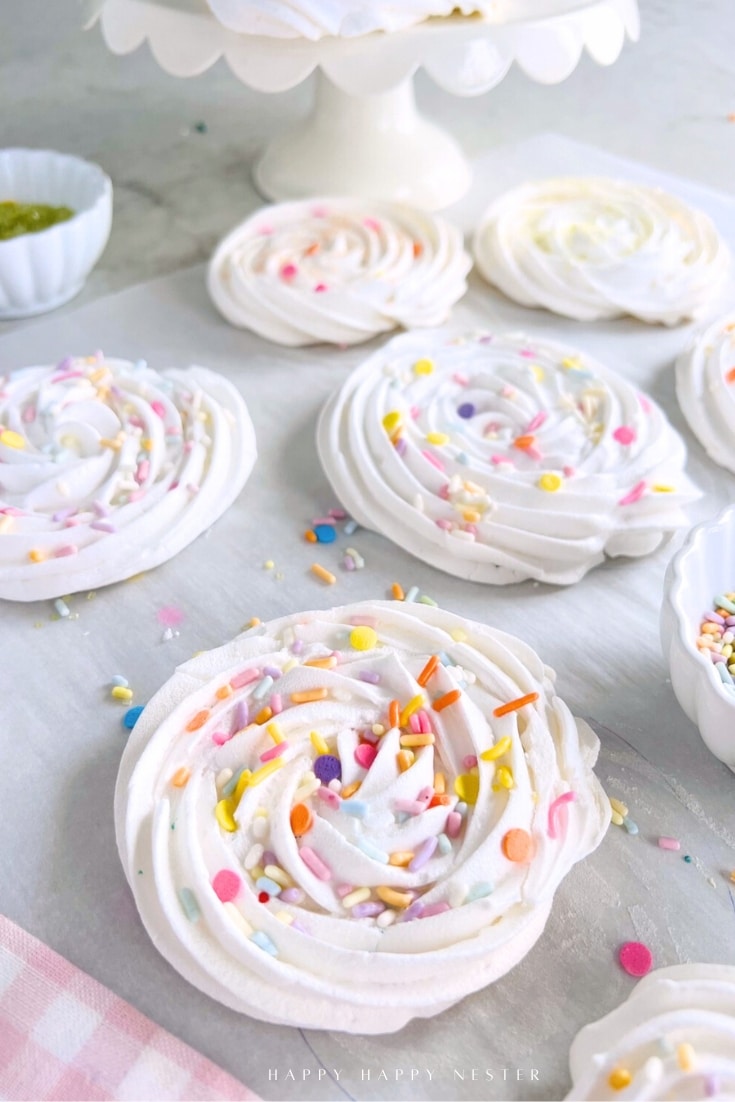 Swirled meringue cookies, made from a delightful meringue cookies recipe, are topped with colorful sprinkles and arranged on parchment. A cake stand and small bowls of extra sprinkles complete the cheerful scene.