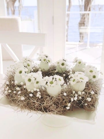 A decorative spring flower wreath DIY centerpiece made of white eggshell vases holding small white flowers and twigs, displayed on a white pedestal dish in a bright room with large windows overlooking water.