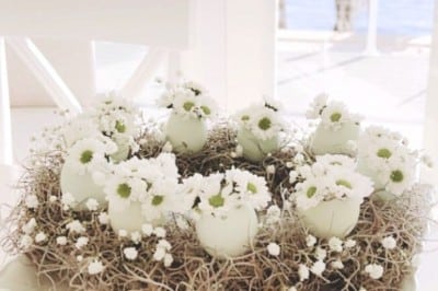 A decorative spring flower wreath DIY centerpiece made of white eggshell vases holding small white flowers and twigs, displayed on a white pedestal dish in a bright room with large windows overlooking water.
