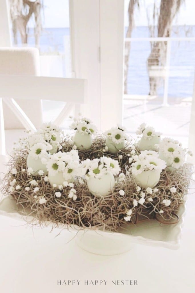 A decorative spring flower wreath DIY centerpiece made of white eggshell vases holding small white flowers and twigs, displayed on a white pedestal dish in a bright room with large windows overlooking water.