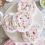 Swirled meringue cookies decorated with colorful sprinkles are arranged on a white cake stand, with more meringues and sprinkles scattered around on a marble surface and a pink gingham cloth nearby.