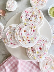 Swirled meringue cookies decorated with colorful sprinkles are arranged on a white cake stand, with more meringues and sprinkles scattered around on a marble surface and a pink gingham cloth nearby.