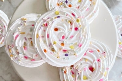 Swirled meringue cookies decorated with colorful sprinkles are arranged on a white cake stand, with more meringues and sprinkles scattered around on a marble surface and a pink gingham cloth nearby.