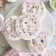 Swirled meringue cookies decorated with colorful sprinkles are arranged on a white cake stand, with more meringues and sprinkles scattered around on a marble surface and a pink gingham cloth nearby.