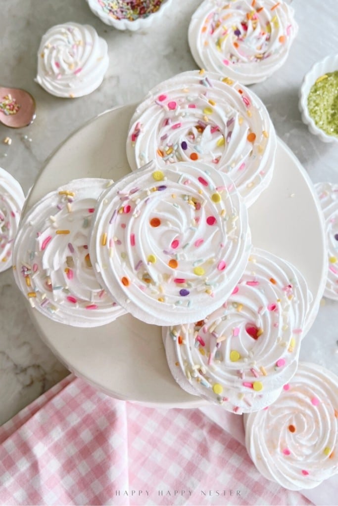 Swirled white meringue cookies topped with colorful sprinkles are arranged on a cake stand, with more cookies and sprinkles scattered around on a marble surface and a pink checkered cloth in the corner.