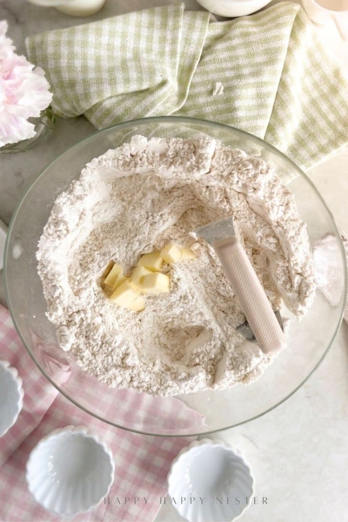 A glass bowl containing flour and small butter cubes with a pastry blender rests on the flour. Surrounding the bowl are empty white ramekins, a pink checkered cloth, and a green checkered cloth.