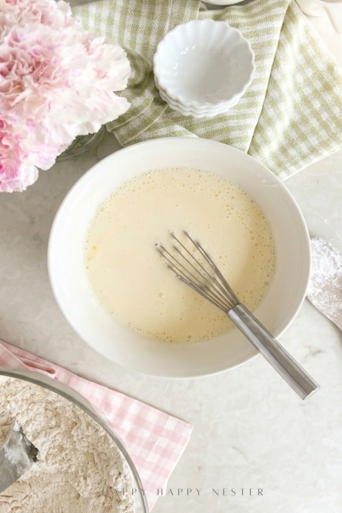 A bowl of batter with a whisk inside sits on a light countertop, surrounded by a bowl of flour, a small dish, a green cloth, and pink flowers.