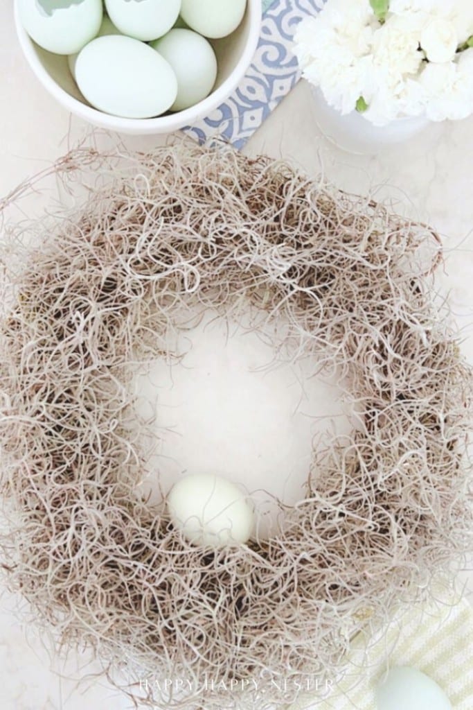 A wreath made of dried twigs and moss with a light green egg inside, placed on a light surface. Nearby are a bowl of pale blue-green eggs and a vase of white flowers.