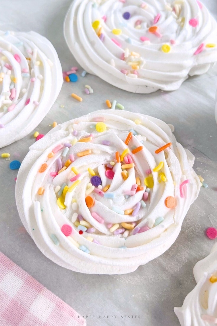 Swirled white meringue cookies, made from an easy meringue cookies recipe, are topped with colorful sprinkles and rest on a light surface, with a pink and white checkered cloth partially visible in the corner.