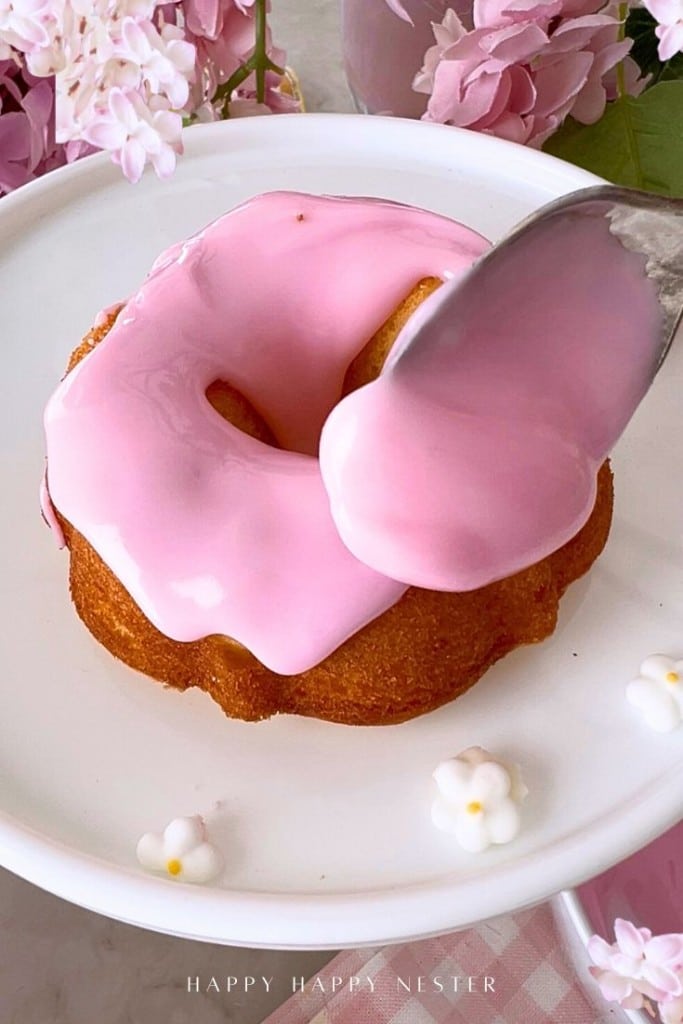 A close-up of a donut on a white plate, being topped with glossy pink icing from a spoon. Pink flowers are in the background, and small white flower decorations are on the plate.