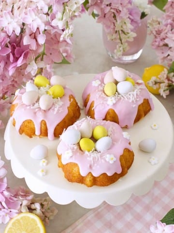 Three mini bundt cakes with pink icing are topped with pastel candy eggs and small white flowers, displayed on a white cake stand surrounded by pink flowers and a glass of pink drink&mdash;perfect for your Easter bundt cake recipe inspiration.
