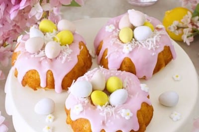 Three mini bundt cakes with pink icing are topped with pastel candy eggs and small white flowers, displayed on a white cake stand surrounded by pink flowers and a glass of pink drink—perfect for your Easter bundt cake recipe inspiration.